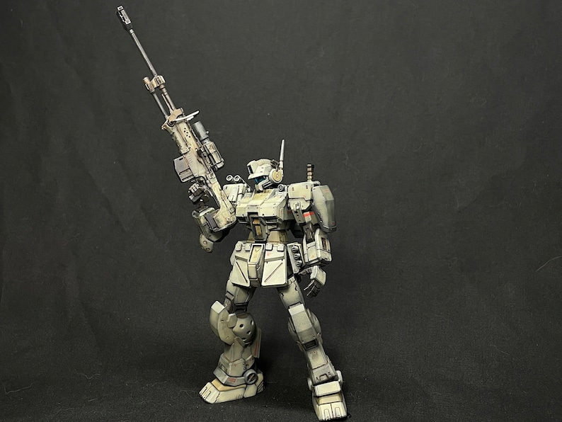 May include: A white and gray robot action figure holding a large rifle. The robot has a helmet with a visor and a detailed design. The figure is posed in a standing position with the rifle raised.