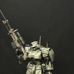 May include: A white and gray robot with a large rifle in its right hand. The robot is standing on a black background.