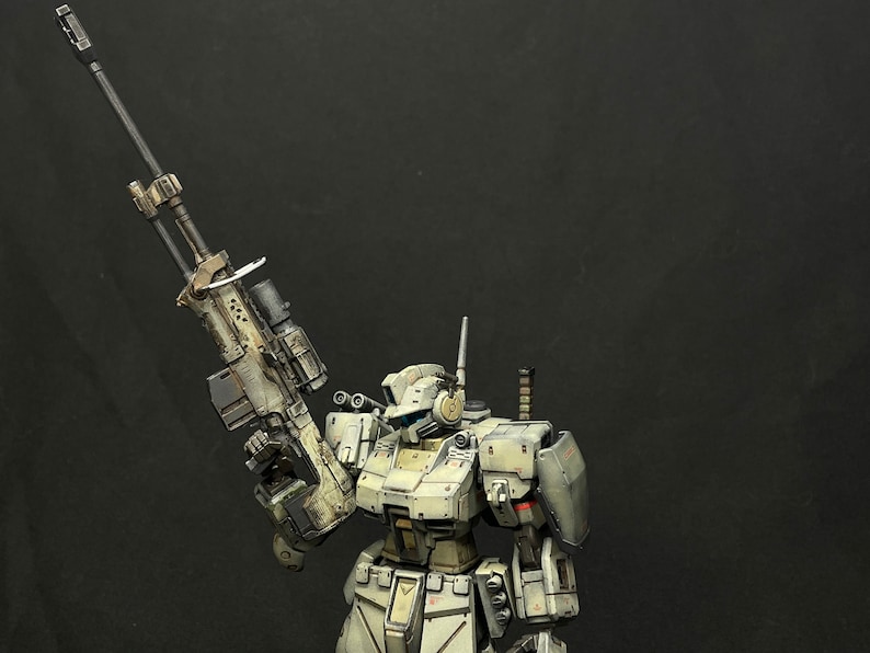 May include: A white and gray robot with a large rifle in its right hand. The robot is standing on a black background.