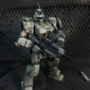 May include: A white and gray robot action figure with a black gun. The robot is standing on a gray surface.