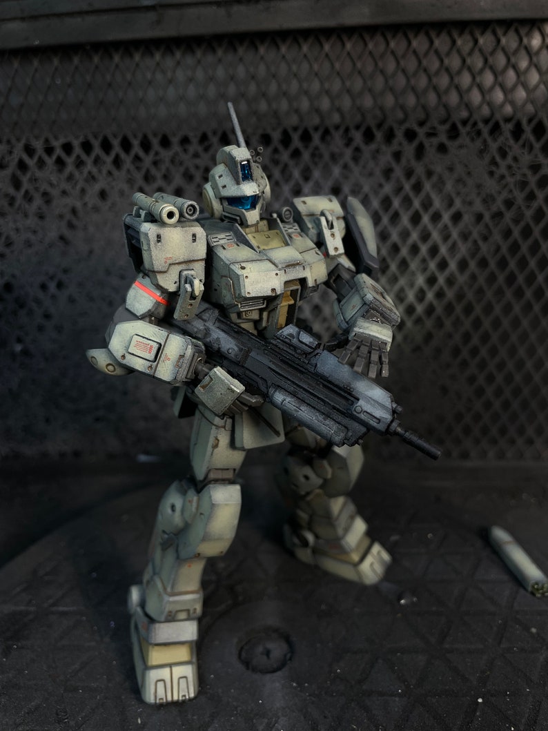 May include: A white and gray robot action figure with a black gun. The robot is standing on a gray surface.