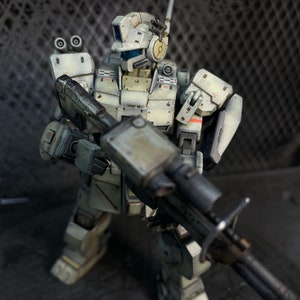 May include: A white and gray robot action figure with a large gun. The robot has a helmet with a visor and a large antenna. The robot is standing on a dark gray surface.