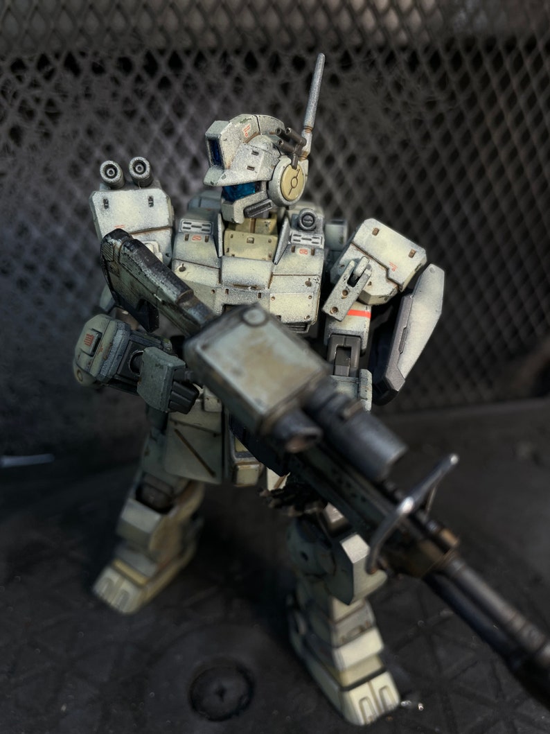May include: A white and gray robot action figure with a large gun. The robot has a helmet with a visor and a large antenna. The robot is standing on a dark gray surface.