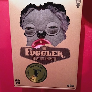 Op de afbeelding: Een bruine kartonnen doos met een uitsparing van een grijs monstergezicht. Het monster heeft grote, slaperige ogen, een zwarte neus en een roze mond. Op de doos staat "Fugglers Funny Ugly Monster" en bevat een gouden sticker.