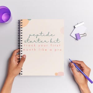 May include: A spiral-bound notebook with the text "peptide starter kit" and "track your first month like a pro". A purple pen and a purple cup are also visible. The notebook is held by two hands.
