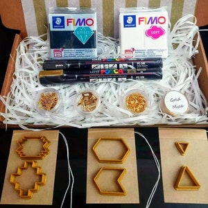 May include: A craft supply kit containing two blocks of polymer clay, one black and one white, labelled "FIMO Effect" and "FIMO Soft". The kit also includes three black metallic markers, two gold-coloured markers, two small containers of gold-coloured jump rings, a small container of gold-coloured beads, a small container of gold mica powder, and three cookie cutters in the shapes of a hexagon, a chevron, and a triangle.