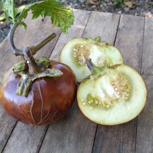 May include: Three eggplants, one whole and two halved, on a wooden surface. The whole eggplant is brown with a green stem. The halved eggplants are light green with white seeds.