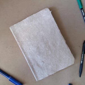 May include: A rectangular notebook with a textured, light beige cover. The notebook is surrounded by several writing instruments, including a green permanent marker, a black ballpoint pen, and two blue pens, all arranged on a flat surface.