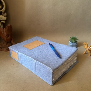 May include: A rectangular, light blue, textured fabric-covered journal with a thick, deckled-edge paper block. The cover features two plain, light brown rectangular patches, one on the front and one on the spine. A blue ballpoint pen rests on top.