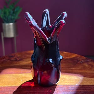 Peut inclure: Un vase en verre rouge foncé avec une forme organique unique. Le vase a un sommet évasé avec plusieurs pointes et une base arrondie. Le verre est translucide, laissant passer la lumière. Il est posé sur une table en bois.