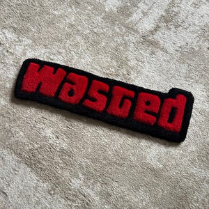 Könnte beinhalten: Ein rechteckiger getufteter Teppich mit dem Wort Wasted in kräftigen roten Buchstaben und einem dicken schwarzen Rand. Der Text hat eine erhabene, strukturierte Floroptik, die einen deutlichen Kontrast zwischen der leuchtend roten Schrift und dem schwarzen Umriss erzeugt.