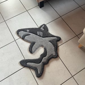 Op de afbeelding: Een grijs en zwart tapijt in de vorm van een haai. Het tapijt heeft een getextureerd, zacht oppervlak en een gelaagd ontwerp, met donkerder grijs dat de randen accentueert. Het ligt op een betegelde vloer.