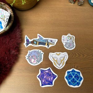 May include: A collection of colorful, die-cut stickers featuring various designs. The stickers include a shark-shaped design with mechanical details, a futuristic tank, and several abstract geometric shapes in shades of blue, purple, and white.