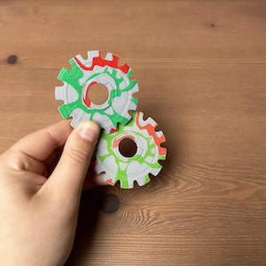 Puede incluir: Dos objetos grises en forma de engranaje con detalles rojos, verdes y naranjas. Los engranajes son sostenidos por una mano sobre un fondo de madera. Los engranajes tienen un agujero circular en el centro y están decorados con un patrón veteado.