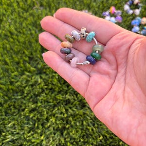 Puede incluir: Un anillo circular de metal plateado que presenta una colección de pequeñas cuentas de piedra lisas y multicolores en tonos turquesa, verde, azul, rosa, marrón y una cuenta blanca con puntos negros, dispuestas alrededor del aro.