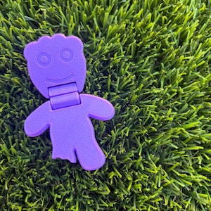 May include: A purple articulated figure shaped like a gummy candy with a textured surface, featuring a rounded head with two circular eyes and a curved mouth, a hinged neck section, and a body with two arms and two legs, one of which is partially missing.