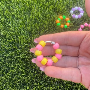 May include: A circular hoop earring featuring alternating bright yellow and hot pink plastic pony beads. The hoop is made of a thin silver-toned metal wire with a small clasp mechanism visible at the top, creating a vibrant and colorful accessory.