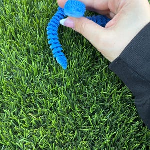 Pode incluir: Um brinquedo de cobra articulado azul brilhante com um corpo texturizado e segmentado e uma cabeça detalhada. O design flexível impresso em 3D apresenta uma série de elos conectados que permitem que o brinquedo se dobre e se mova, exibindo uma cor sólida vibrante.