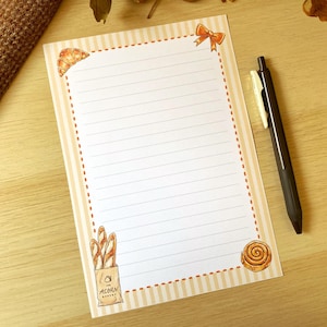 May include: A lined notepad with a bakery theme. The notepad has a striped border with orange dotted lines. Illustrations of baked goods, including a croissant, baguettes, and a cinnamon roll, are featured. A black pen rests on the right side.