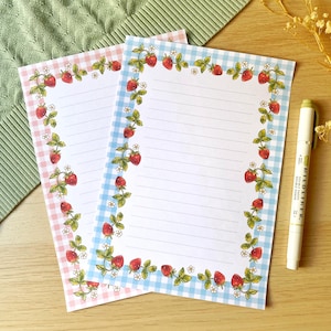 May include: Two lined notepads with strawberry and gingham borders. One notepad has a pink gingham border, the other has a blue gingham border. A white pen with green accents rests on the wooden surface.