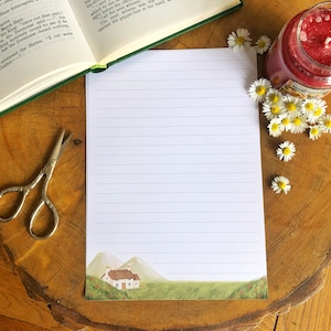 May include: Lined writing paper featuring a watercolour illustration of a cottage and mountains. A red candle, daisies, scissors, and a book are arranged on a wooden surface, creating a rustic, homely feel.