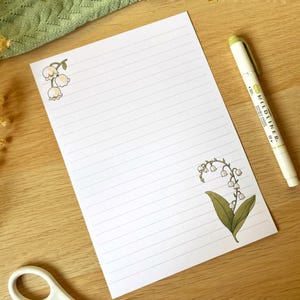 May include: A white sheet of lined paper with two illustrations of lily of the valley flowers. The paper is on a wooden surface with a yellow highlighter pen.