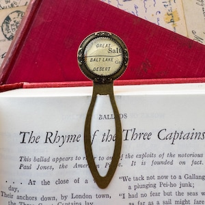 May include: A brass bookmark with a round glass charm featuring a vintage map of the Great Salt Lake Desert. The bookmark is shaped like an oval and has the word "BALLADS" printed on it.