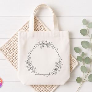 May include: Cream-colored tote bag with a black floral geometric design. The bag has a rectangular shape with a handle. The design is a black floral wreath with a geometric shape in the center. The bag is sitting on a woven mat.