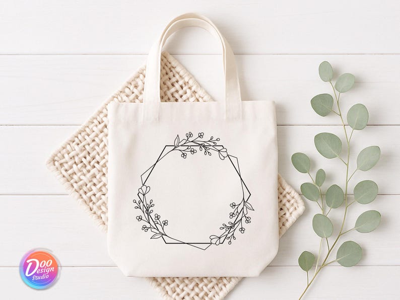 May include: Cream-colored tote bag with a black floral geometric design. The bag has a rectangular shape with a handle. The design is a black floral wreath with a geometric shape in the center. The bag is sitting on a woven mat.