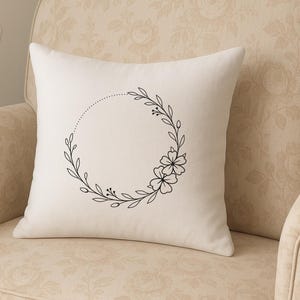 May include: A cream-colored pillow with a black floral wreath design. The wreath is composed of leaves, berries, and flowers, forming a circle with a dotted line. The pillow is on a beige floral patterned chair.