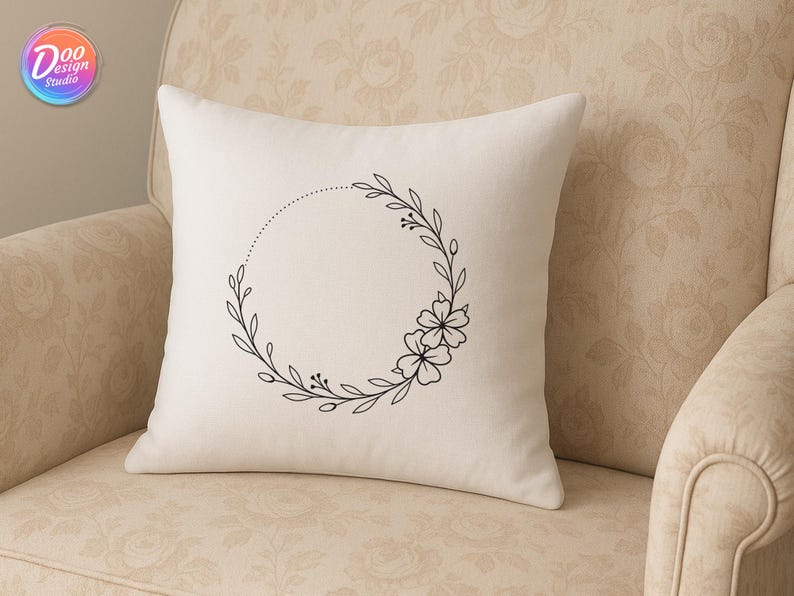 May include: A cream-colored pillow with a black floral wreath design. The wreath is composed of leaves, berries, and flowers, forming a circle with a dotted line. The pillow is on a beige floral patterned chair.