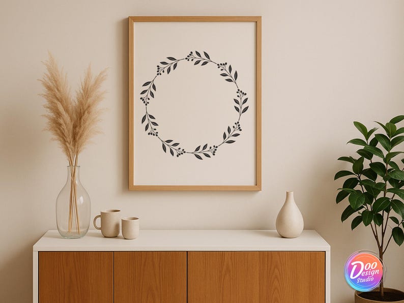 May include: A framed print with a black botanical wreath design hangs above a white cabinet. A clear glass vase with dried pampas grass, two beige mugs, and a white vase are on the cabinet. A potted plant is on the right.