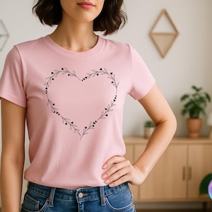 May include: A light pink t-shirt featuring a black heart-shaped design made of leaves and small hearts. The short-sleeved shirt is made of soft material and is suitable for casual wear. The design is centered on the chest.