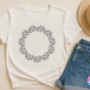 May include: White t-shirt featuring a black floral wreath design. The wreath is composed of leaves and small berries, forming a circular shape. A straw hat and denim shorts are also visible.