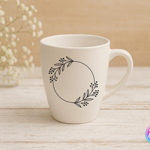 May include: A white ceramic mug with a black floral wreath design. The wreath is a circular arrangement of leaves and berries. The mug has a curved handle and is set against a light wooden background.