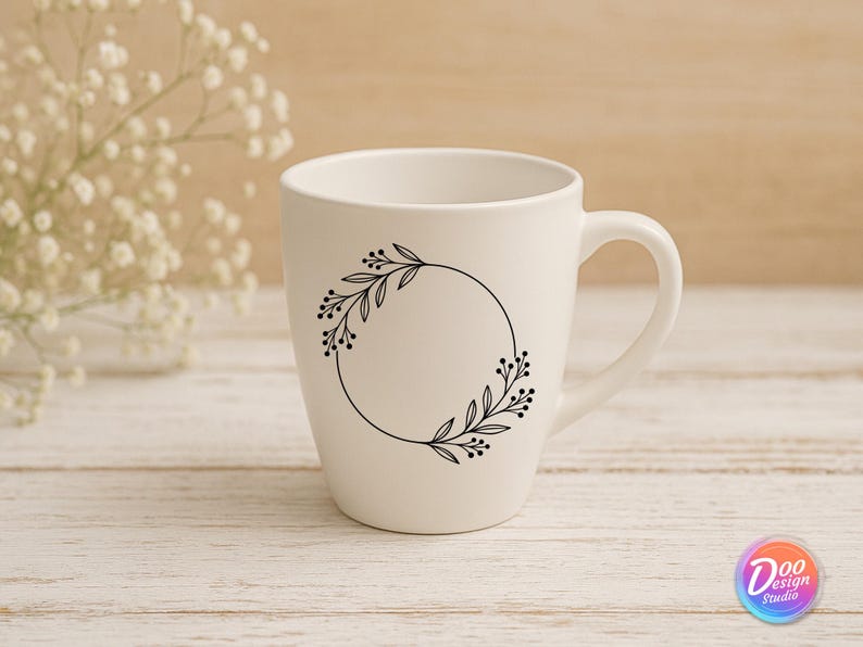 May include: A white ceramic mug with a black floral wreath design. The wreath is a circular arrangement of leaves and berries. The mug has a curved handle and is set against a light wooden background.