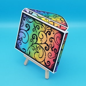 Pode incluir: Uma caixa com cores de arco-íris com um design floral preto e branco. A caixa está em um pequeno cavalete de madeira.