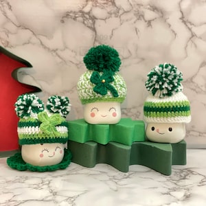 May include: Three ceramic marshmallow figures wearing green and white crochet hats with pom-poms and shamrock accents. The figures are displayed on a green star-shaped platform against a marble background.