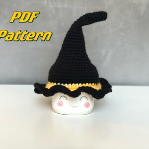 Könnte beinhalten: Ein schwarzer Häkelhut mit gelbem Rand, der auf einer weißen Tasse mit einem lächelnden Gesicht sitzt. Der Text "PDF Pattern" befindet sich in der oberen linken Ecke des Bildes.