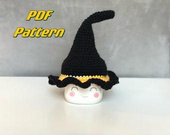 May include: A black crocheted witch hat with a yellow brim, sitting on a white mug with a smiling face. The text "PDF Pattern" is in the top left corner of the image.
