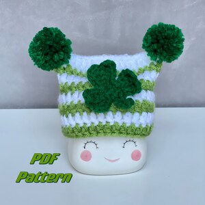 May include: A white ceramic marshmallow with a smiling face wearing a green, white, and green striped crochet hat with green pom-poms and a shamrock. The text "PDF Pattern" is displayed in green.