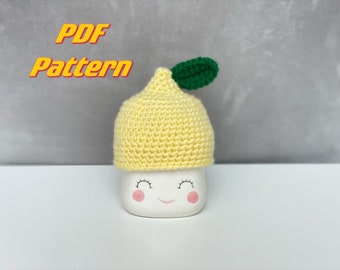 May include: A crocheted lemon-shaped hat with a green leaf accent. The hat is yellow and sits atop a white ceramic container with a smiling face. The words "PDF Pattern" are in yellow and orange.