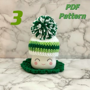 May include: A white crocheted mug cozy with green and white stripes and a white and green pom pom on top. The cozy is on a white mug with a smiling face. The text "PDF Pattern" is in the upper right corner of the image. The number "3" is in the upper left corner of the image.
