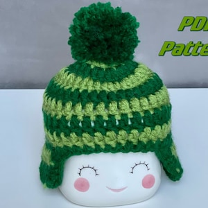 May include: A green and lime green striped crochet hat with a green pom pom on top. The hat has ear flaps and is sitting on a white surface. PDF Pattern.