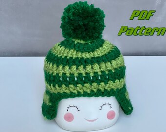 May include: A green and lime green striped crochet hat with a green pom pom on top. The hat has ear flaps and is sitting on a white surface. PDF Pattern.