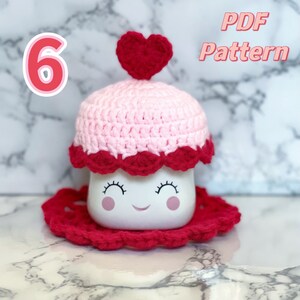 May include: A pink and red crocheted mug cozy with a heart-shaped top. The cozy is designed to fit a mug with a smiling face. The text "PDF Pattern" is visible in the top right corner of the image.