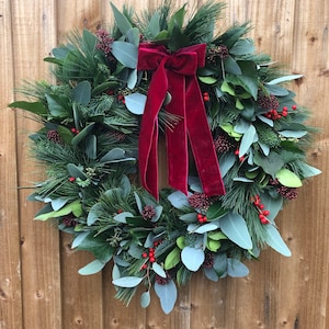 May include: A circular wreath constructed from green pine needles, eucalyptus leaves, and dark green foliage, accented with clusters of small red berries. A large, deep red velvet ribbon with two long, hanging tails is tied at the top center.