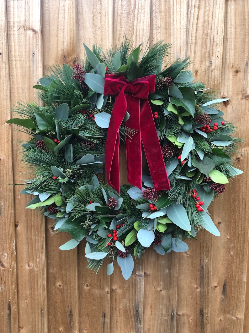 May include: A circular wreath constructed from green pine needles, eucalyptus leaves, and dark green foliage, accented with clusters of small red berries. A large, deep red velvet ribbon with two long, hanging tails is tied at the top center.