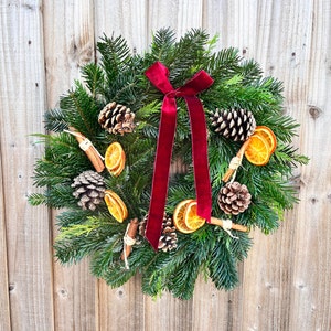 May include: A festive Christmas wreath made of evergreen branches, adorned with pine cones, dried orange slices, and cinnamon sticks. A deep red velvet ribbon is tied in a bow at the top. The wreath is displayed against a wooden background.