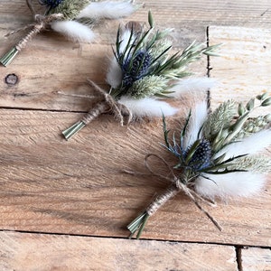 May include: Three small bouquets of dried flowers tied with twine. Each bouquet features blue thistle flowers, white fluffy dried grasses, and green dried grasses.